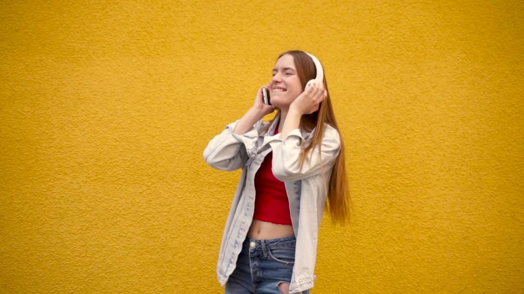 Молодая девушка слушает музыку в наушниках и танцует на фоне желтой стены